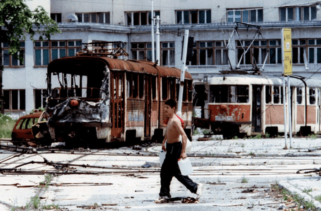 A Bosnian teenager carrying containers of water in front of destroyed trams at Skenderia Square